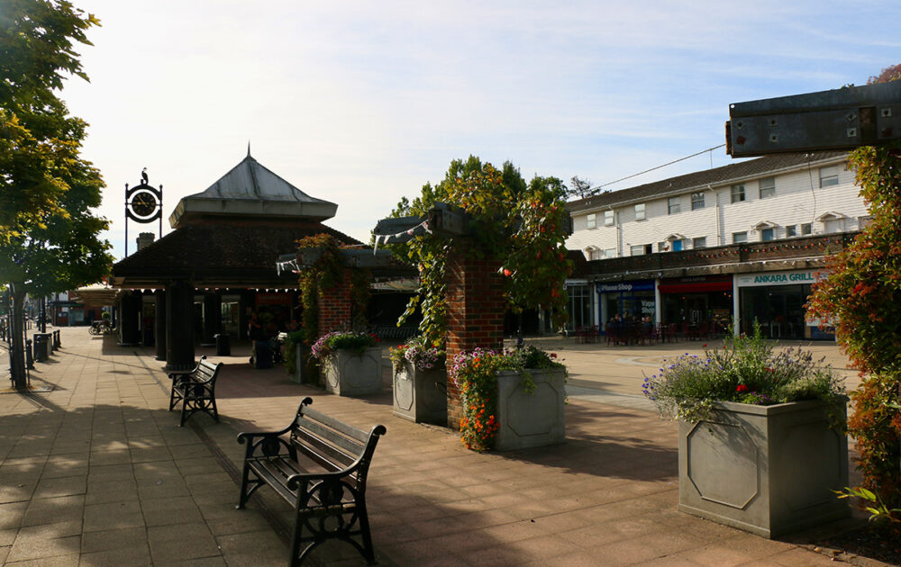 Colour photograph of Stocklund Square in Cranleigh. There are shops, a takeaway, a supermarket and pots overflowing with flowers. The depiction of a crane is visible on top of a large clock.